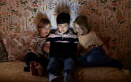 Tre barn tittar på dator i säng. 
