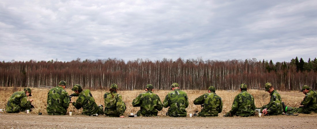 Soldater sitter på rad på ett fält och äter. 