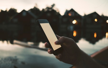 Hand håller i mobiltelefon i kvällsljus med vatten och hus i bakgrunden.