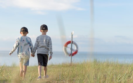 Två barn håller hand på en strand. Livboj och hav i bakgrunden.