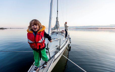 Barn med röd flytväst på en segelbåt. Hav i bakgrunden. 