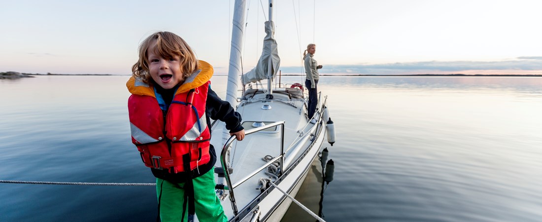Barn med röd flytväst på en segelbåt. Hav i bakgrunden. 