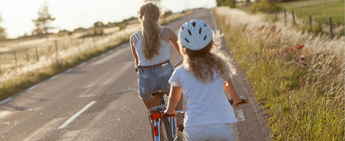 Två flickor som cyklar på en landsväg. Det är sommar och båda flickorna har på sig kortärmat. Den ena flickan använder cykelhjälm. 
