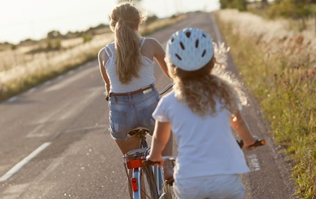 Två flickor som cyklar på en landsväg. Det är sommar och båda flickorna har på sig kortärmat. Den ena flickan använder cykelhjälm. 