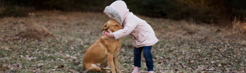 Ett barn i rosa jacka kramar en hund utomhus.