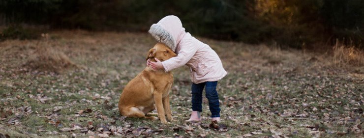 Liten barn i ljus regnjacka kramar hund som sitter ned.