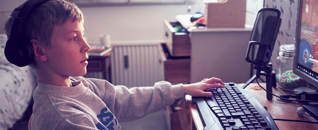 Pojke fotad i profil när han spelar dator vid sitt skrivbord. Pojkens blick är fäst vid skärmen och hans händer vilar på tangentbordet och datormusen. 