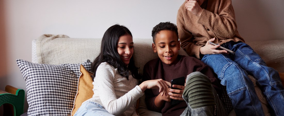 Tre barn sitter i soffan och tittar i en telefon. Barnen skrattar åt något de ser på telefonen. 