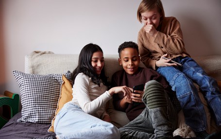 Tre barn sitter i soffan och tittar i en telefon. Barnen skrattar åt något de ser på telefonen. 