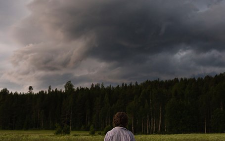 En person står på ett fält vid en skog, himlen är mörk och full av moln