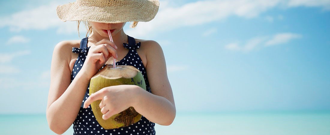 Flicka i solhatt som döljer ansiktet. Flickan dricker ur en kokosnöt med sugrör.