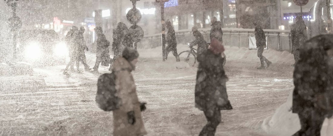 Människor i stadsmiljö går över väg i snöstorm. 