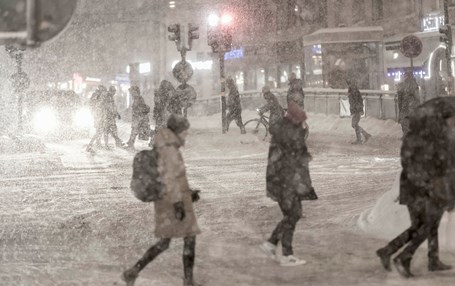 Människor i stadsmiljö går över väg i snöstorm. 