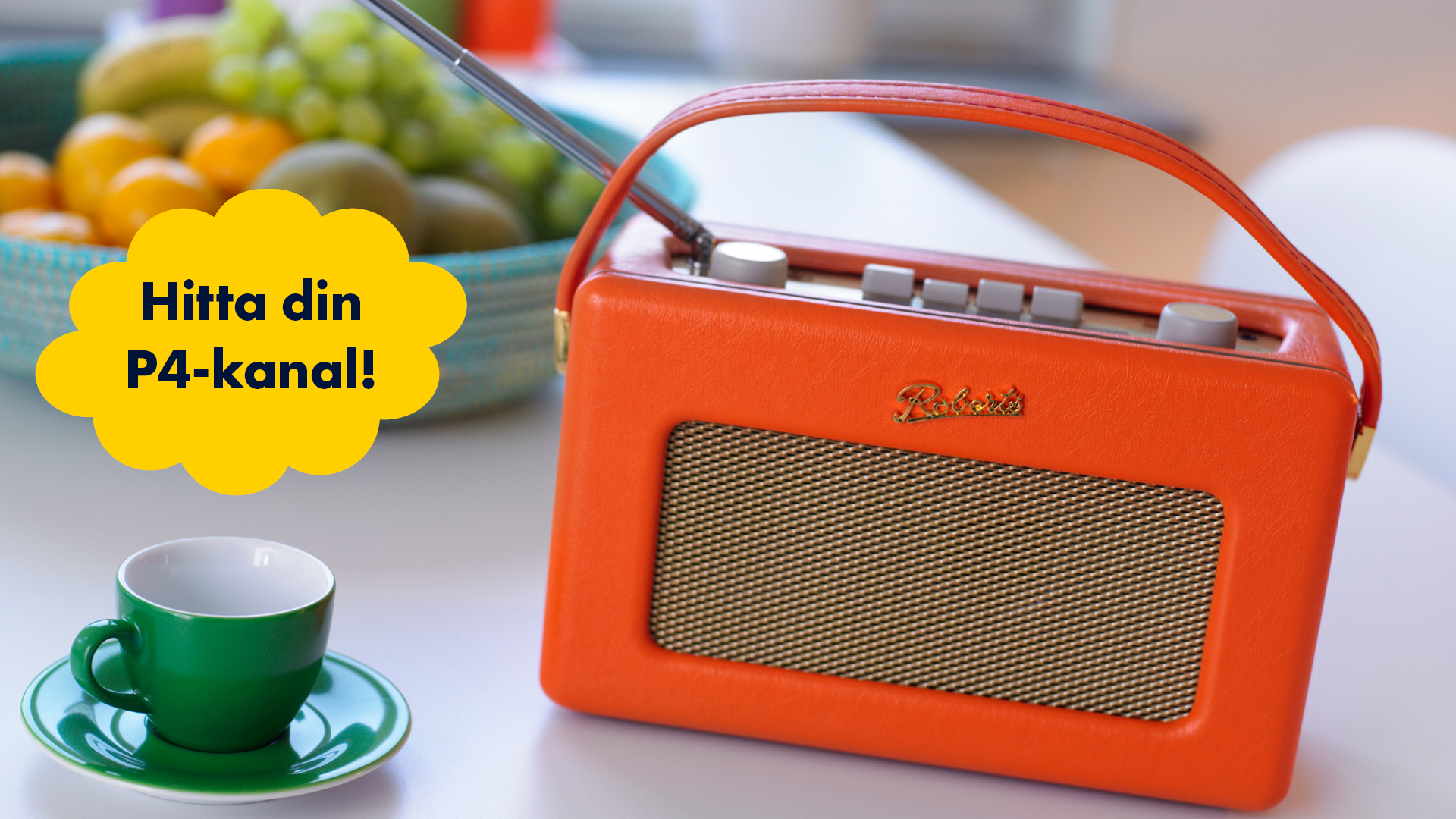 Närbild på en orange radio och en grön kopp på ett vitt bord. I bakgrunden syns en fruktskål.