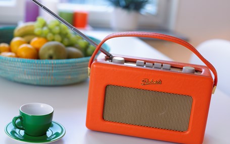 Närbild på en orange radio och en grön kopp på ett vitt bord. I bakgrunden syns en fruktskål.