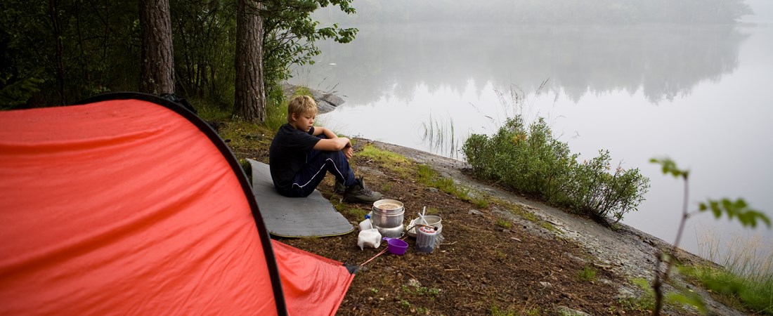 Barn sitter på en klippa vid en sjö. I bakgrunden står ett tält. Bredvid barnet finns ett stormkök uppställt. 