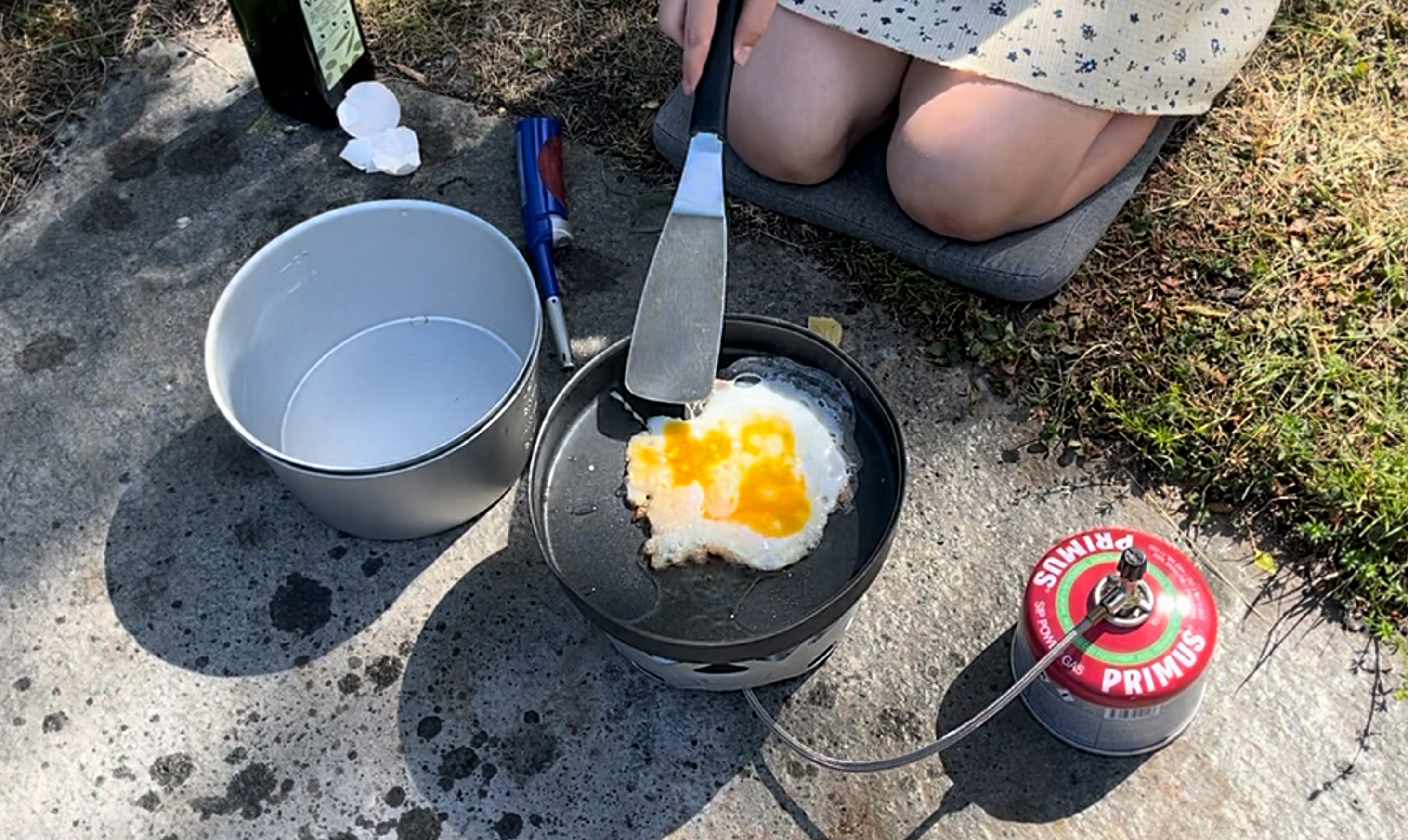 Närbild på ett gasolkök. Ett barn sitter på knä och steker ett ägg i stekpannan.